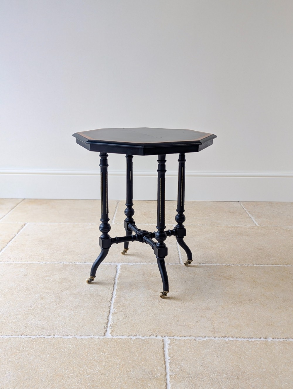 A late Victorian ebonised octagonal occasional table featuring turned legs, gilt highlights, cross stretchers, and brass castors.