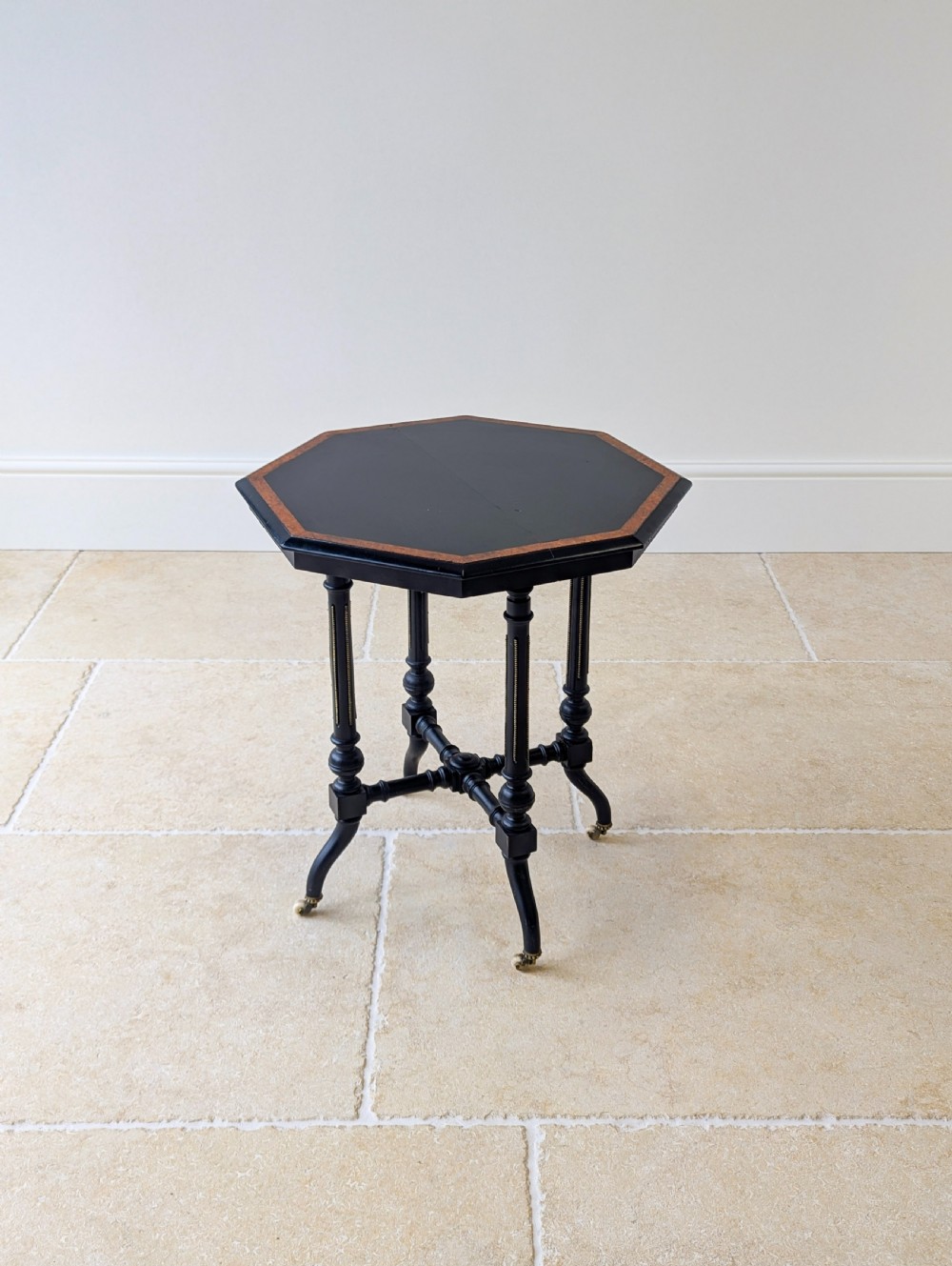A late Victorian ebonised octagonal occasional table featuring turned legs, gilt highlights, cross stretchers, and brass castors. View III