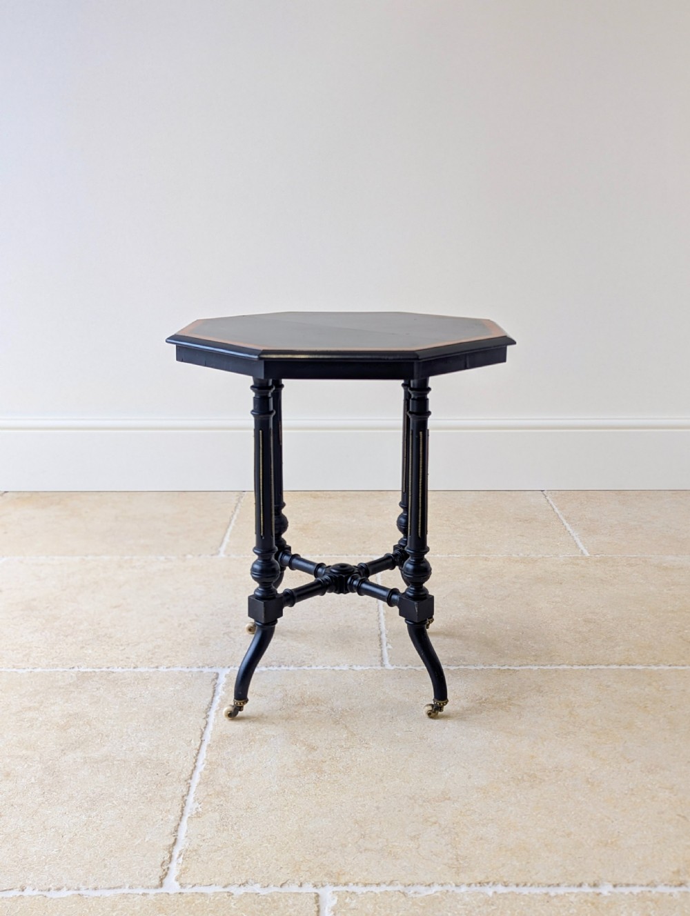 A late Victorian ebonised octagonal occasional table featuring turned legs, gilt highlights, cross stretchers, and brass castors. View V