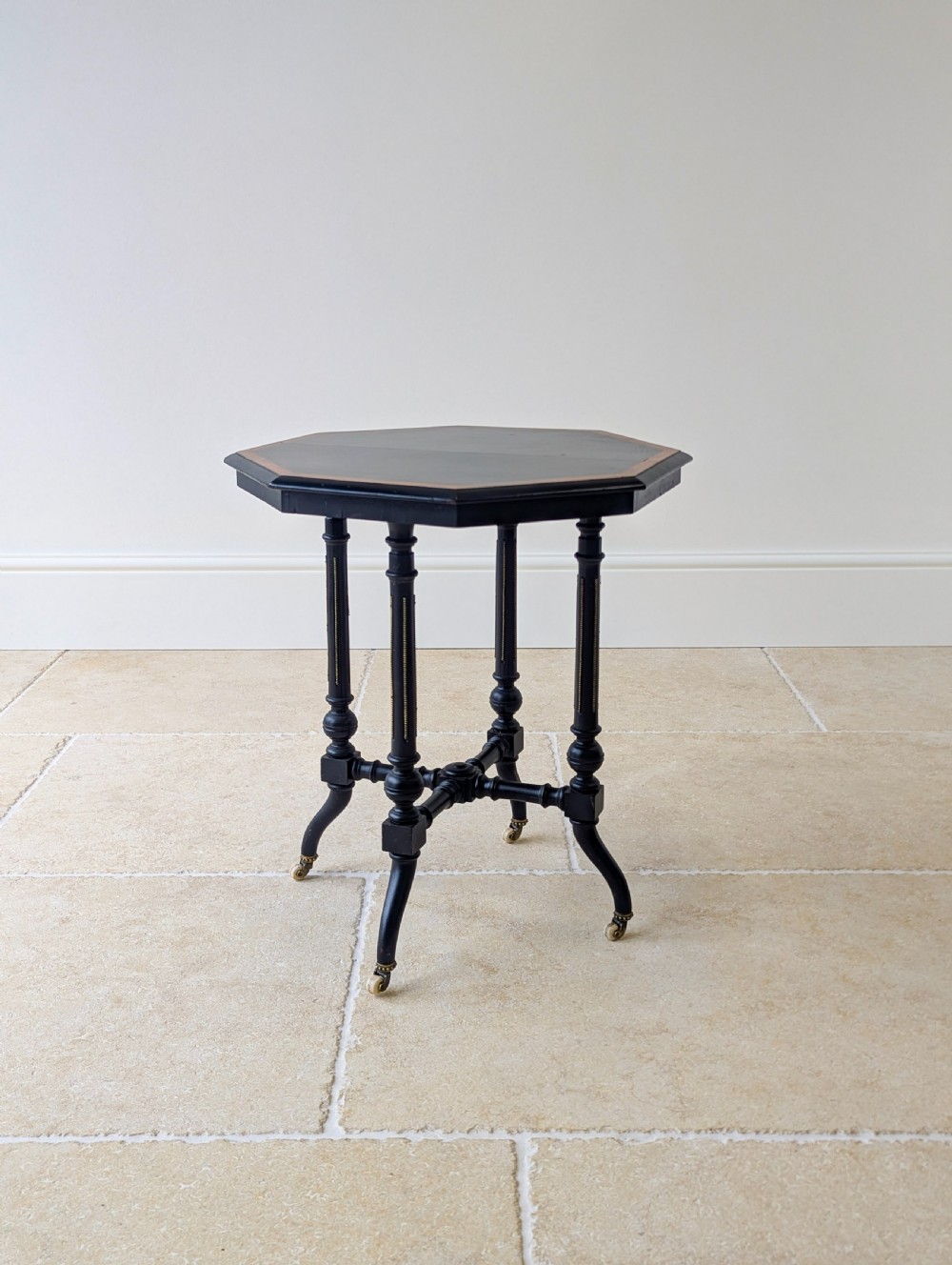 A late Victorian ebonised octagonal occasional table featuring turned legs, gilt highlights, cross stretchers, and brass castors. View VII