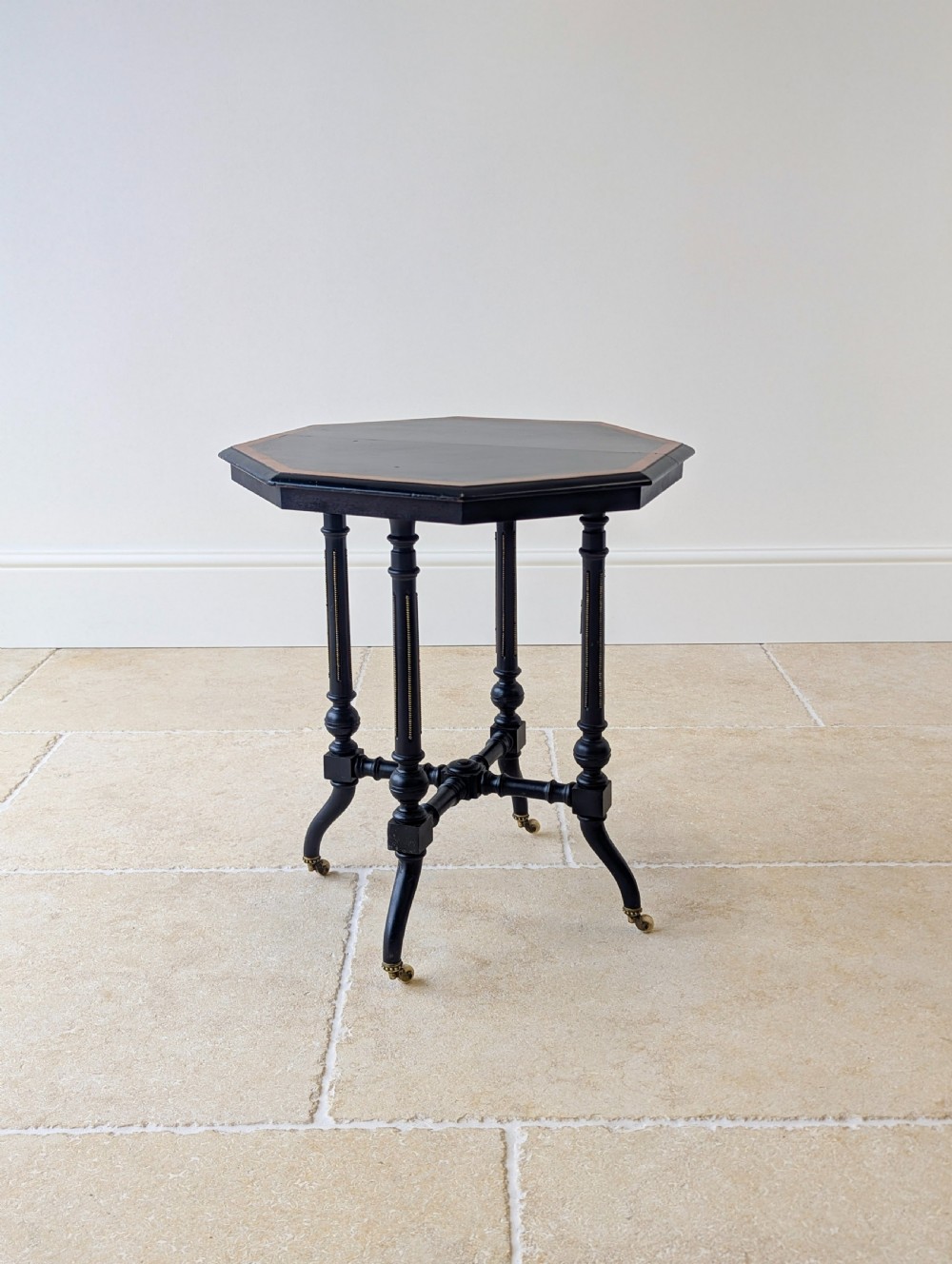A late Victorian ebonised octagonal occasional table featuring turned legs, gilt highlights, cross stretchers, and brass castors. View IX