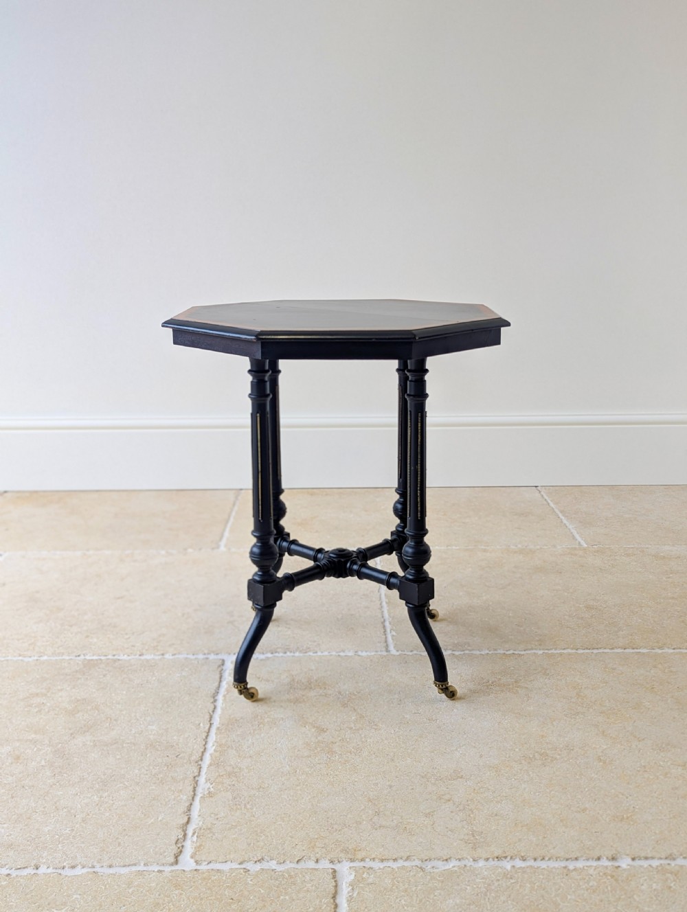 A late Victorian ebonised octagonal occasional table featuring turned legs, gilt highlights, cross stretchers, and brass castors. View XI