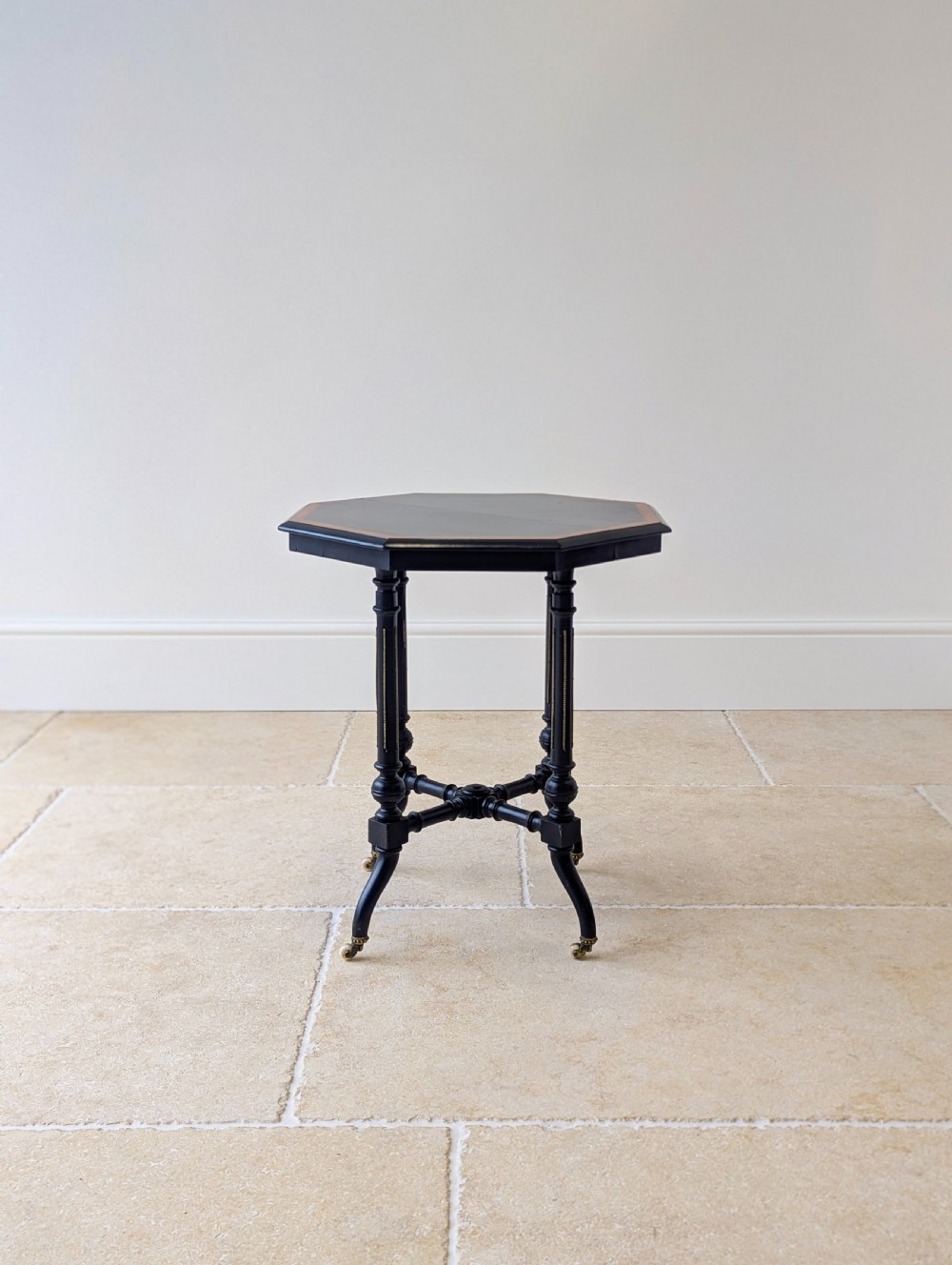 A late Victorian ebonised octagonal occasional table featuring turned legs, gilt highlights, cross stretchers, and brass castors. View XIII