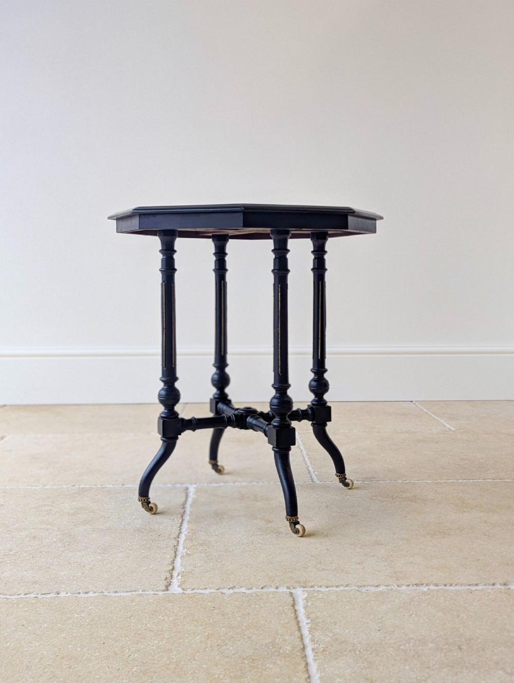 A late Victorian ebonised octagonal occasional table featuring turned legs, gilt highlights, cross stretchers, and brass castors. View XV
