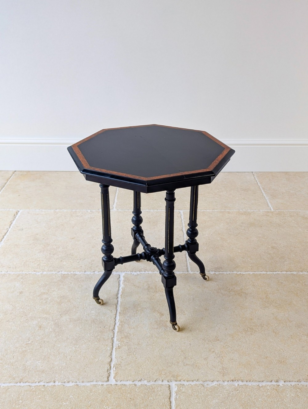 A late Victorian ebonised octagonal occasional table featuring turned legs, gilt highlights, cross stretchers, and brass castors. View XVII