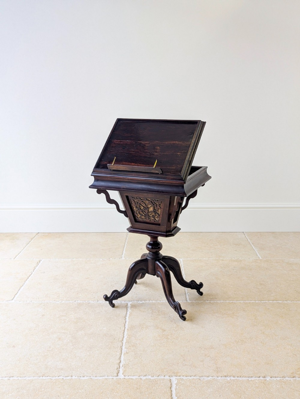 A dark brown rosewood Victorian sewing and reading table featuring an adjustable slanted lid with brass page holders, carved fretwork sides, a turned pedestal column, and four scrolled feet.