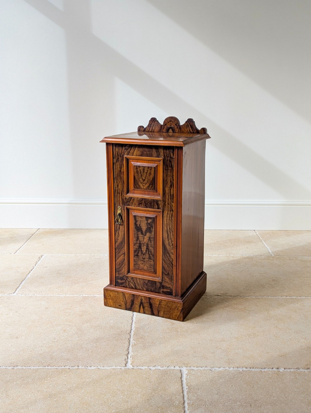 A late Victorian mahogany and exotic wood bedside cupboard featuring a carved top gallery, panelled door with brass drop handle, and a solid plinth base.