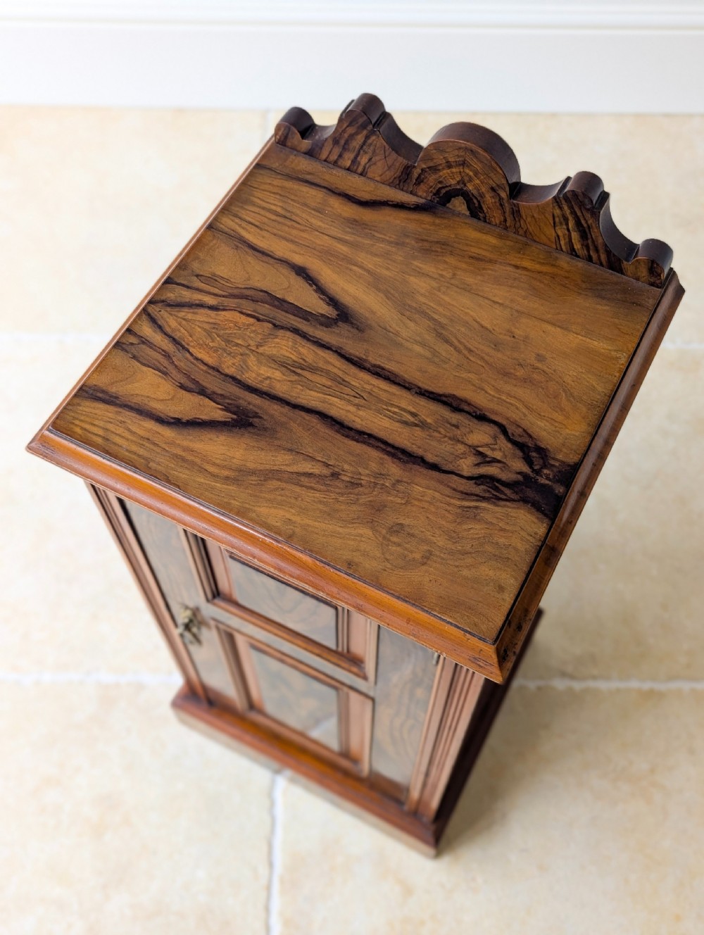 A late Victorian mahogany and exotic wood bedside cupboard featuring a carved top gallery, panelled door with brass drop handle, and a solid plinth base. View II