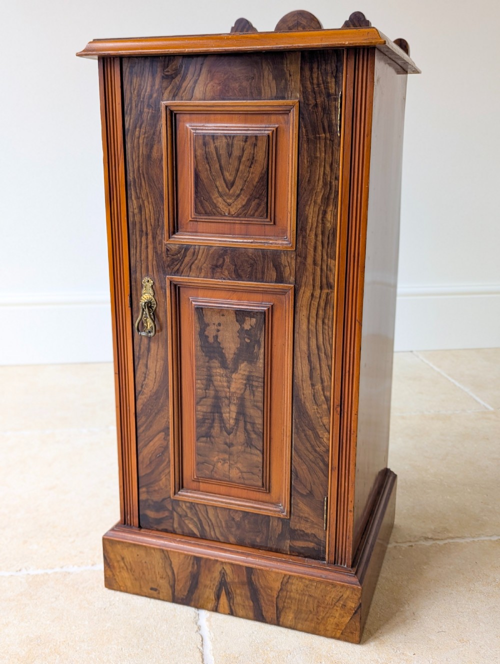 A late Victorian mahogany and exotic wood bedside cupboard featuring a carved top gallery, panelled door with brass drop handle, and a solid plinth base. View IV