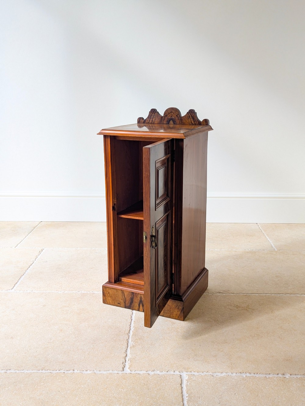 A late Victorian mahogany and exotic wood bedside cupboard featuring a carved top gallery, panelled door with brass drop handle, and a solid plinth base. View V