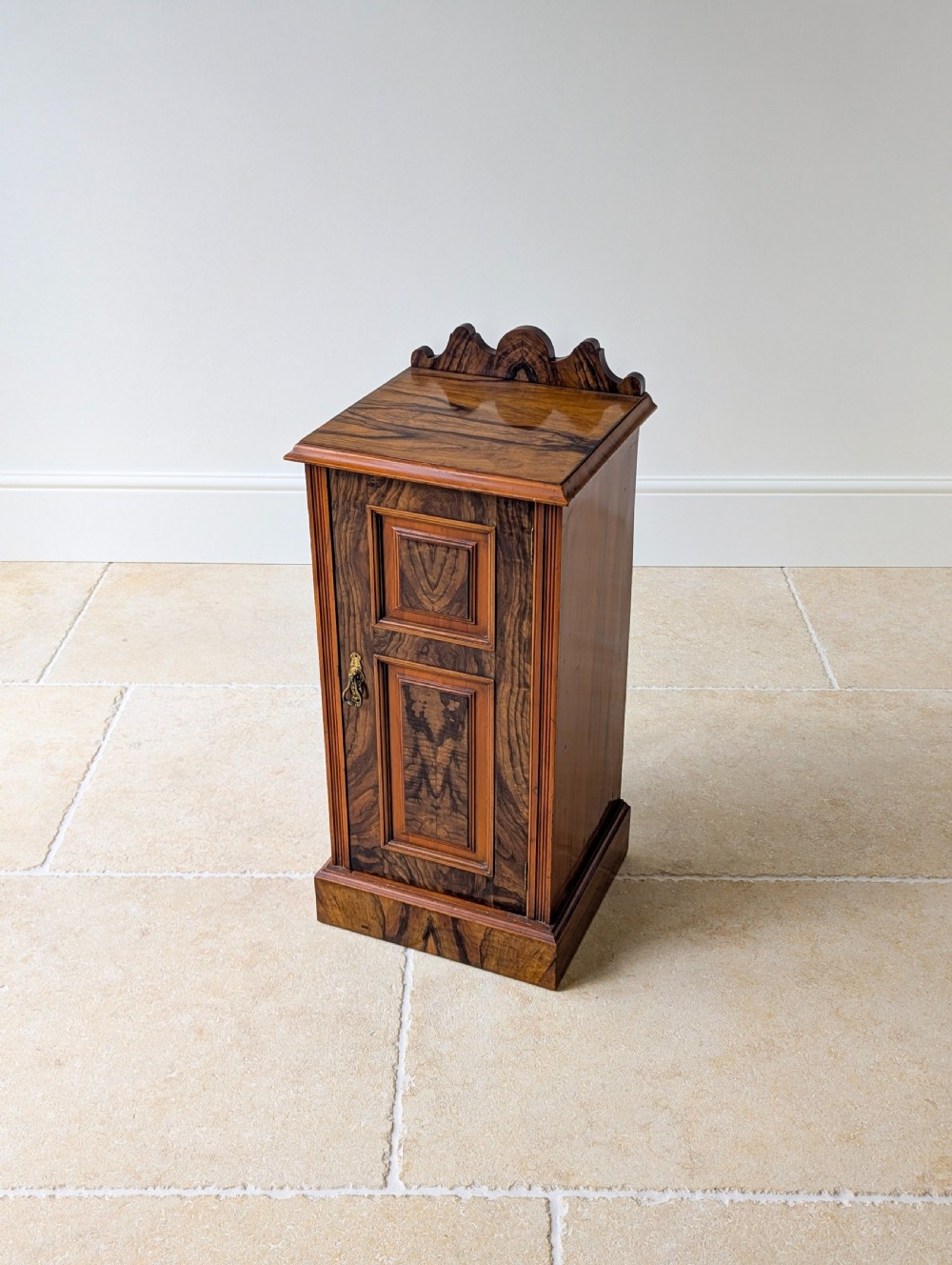 A late Victorian mahogany and exotic wood bedside cupboard featuring a carved top gallery, panelled door with brass drop handle, and a solid plinth base. View VII