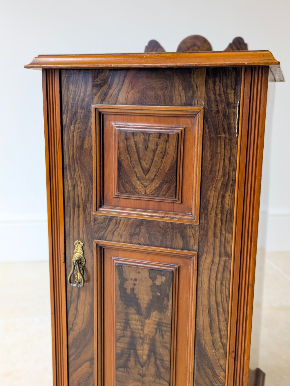 A late Victorian mahogany and exotic wood bedside cupboard featuring a carved top gallery, panelled door with brass drop handle, and a solid plinth base. View X
