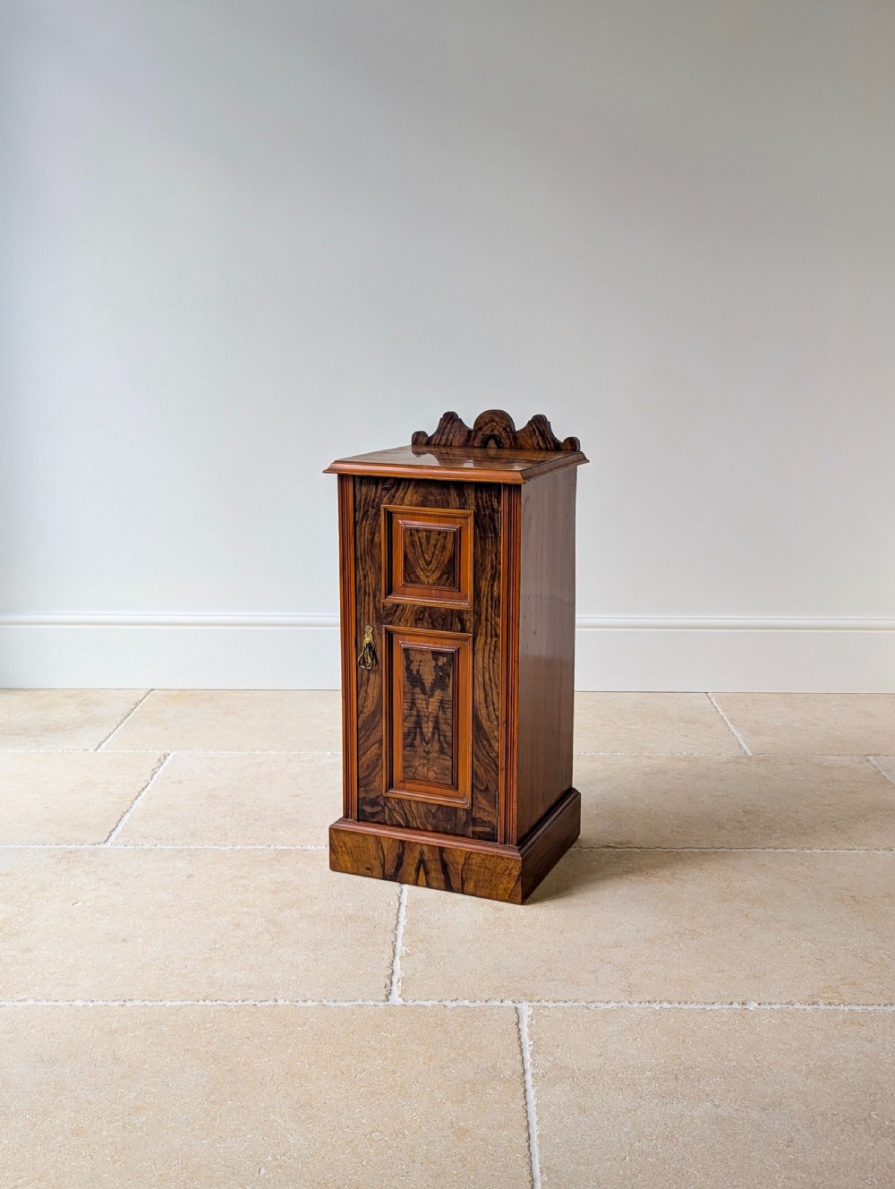 A late Victorian mahogany and exotic wood bedside cupboard featuring a carved top gallery, panelled door with brass drop handle, and a solid plinth base. View XI