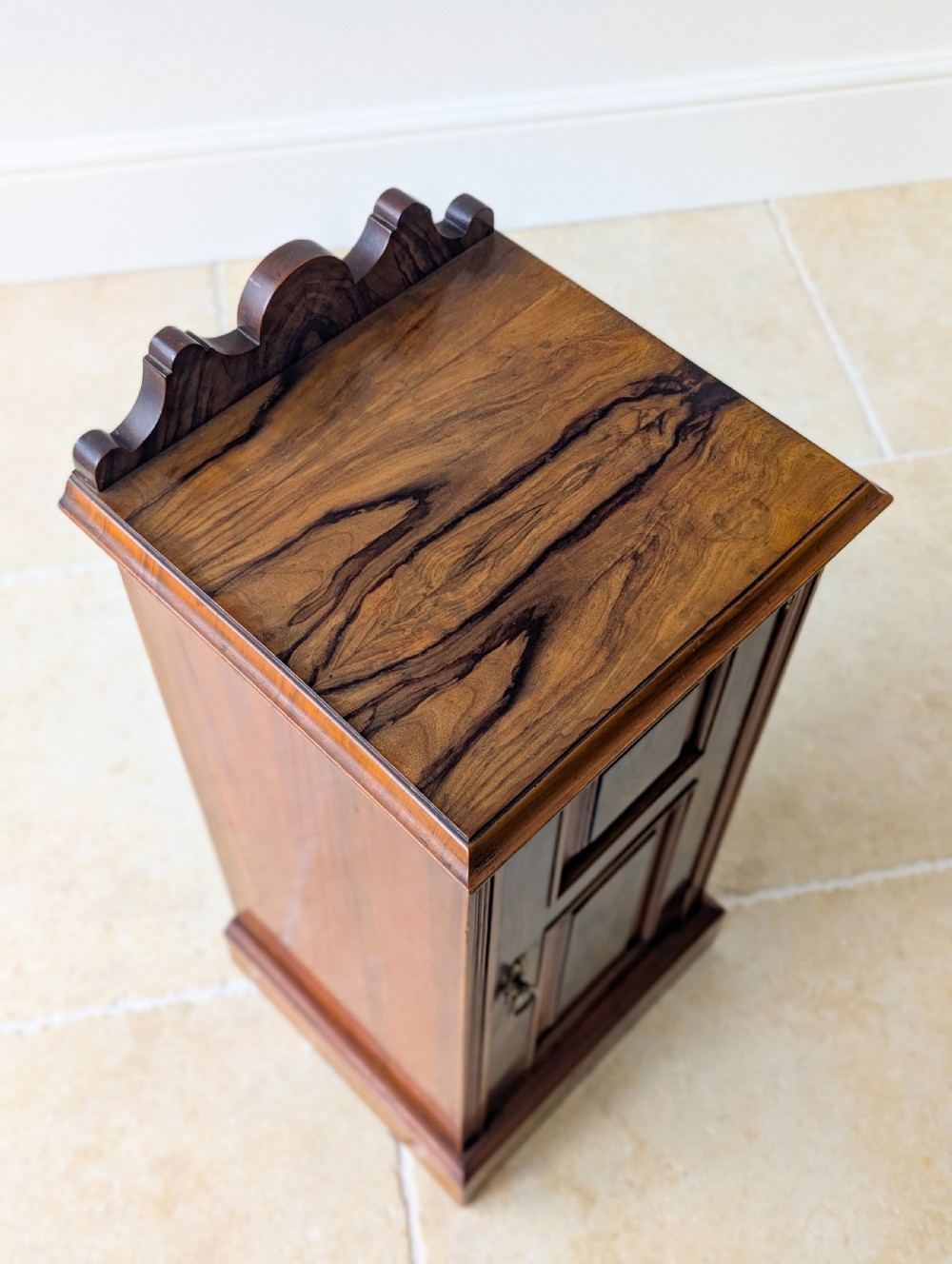 A late Victorian mahogany and exotic wood bedside cupboard featuring a carved top gallery, panelled door with brass drop handle, and a solid plinth base. View XII
