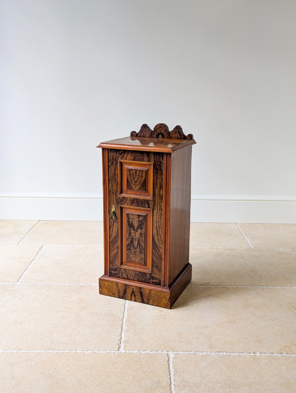 A late Victorian mahogany and exotic wood bedside cupboard featuring a carved top gallery, panelled door with brass drop handle, and a solid plinth base. View XIV