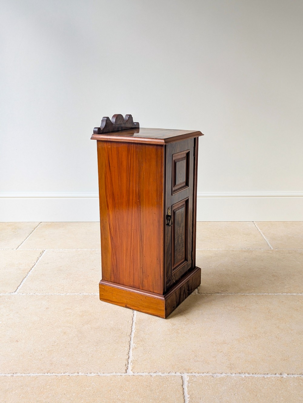 A late Victorian mahogany and exotic wood bedside cupboard featuring a carved top gallery, panelled door with brass drop handle, and a solid plinth base. View XV