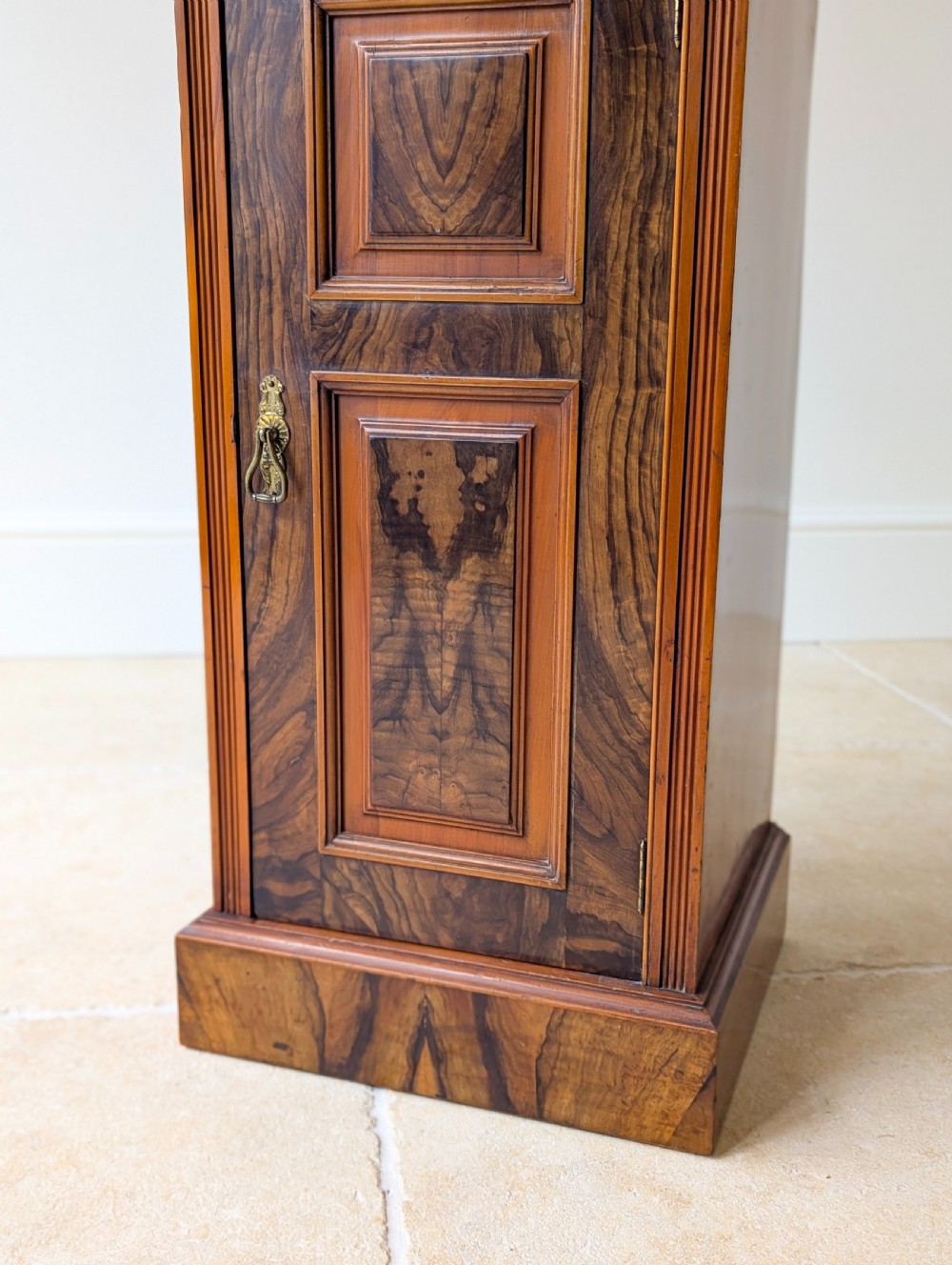 A late Victorian mahogany and exotic wood bedside cupboard featuring a carved top gallery, panelled door with brass drop handle, and a solid plinth base. View XVII