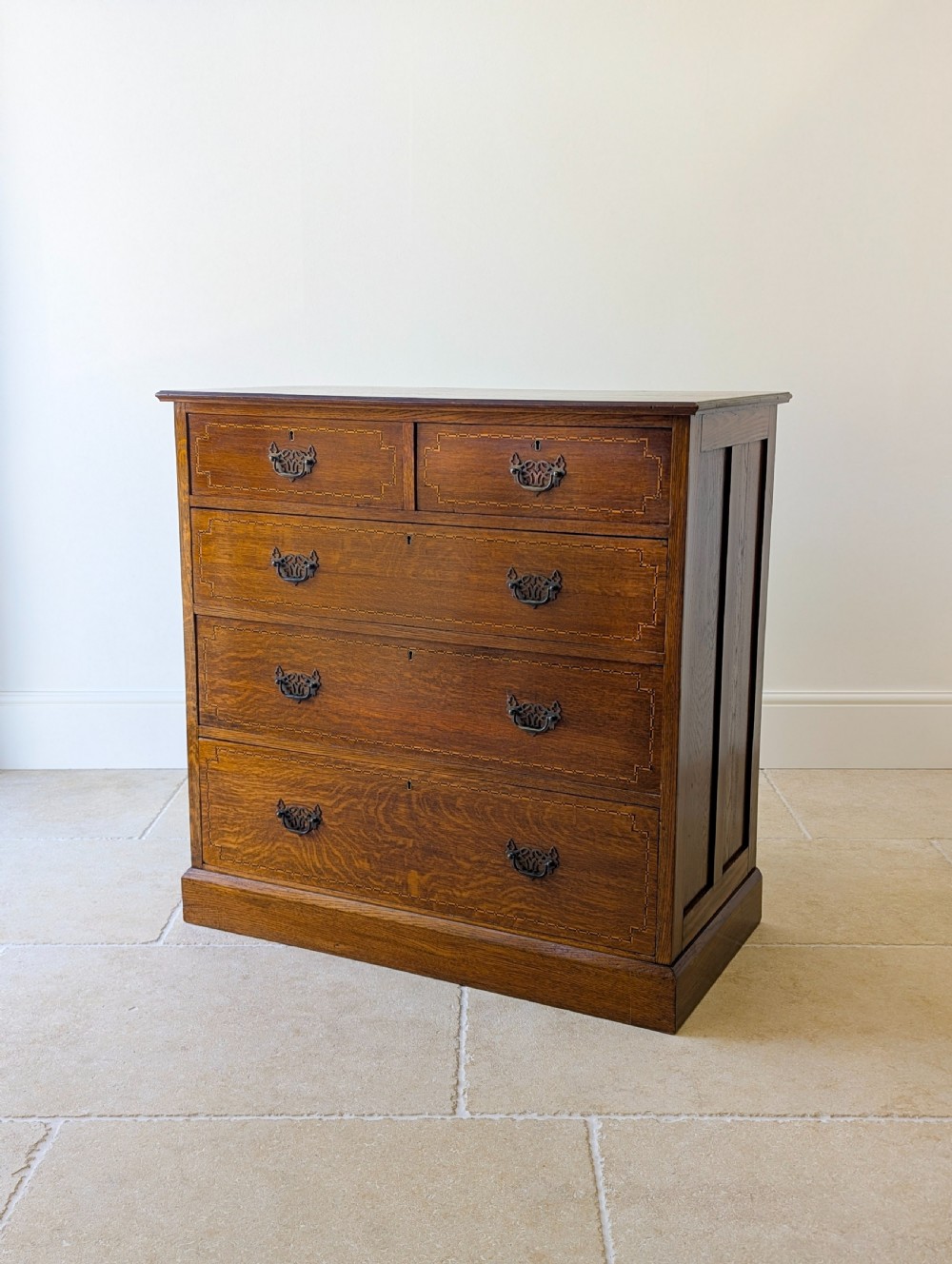 An Edwardian oak chest of drawers featuring a two over three layout with geometric boxwood inlay and dark metal drop handles on a plinth base.