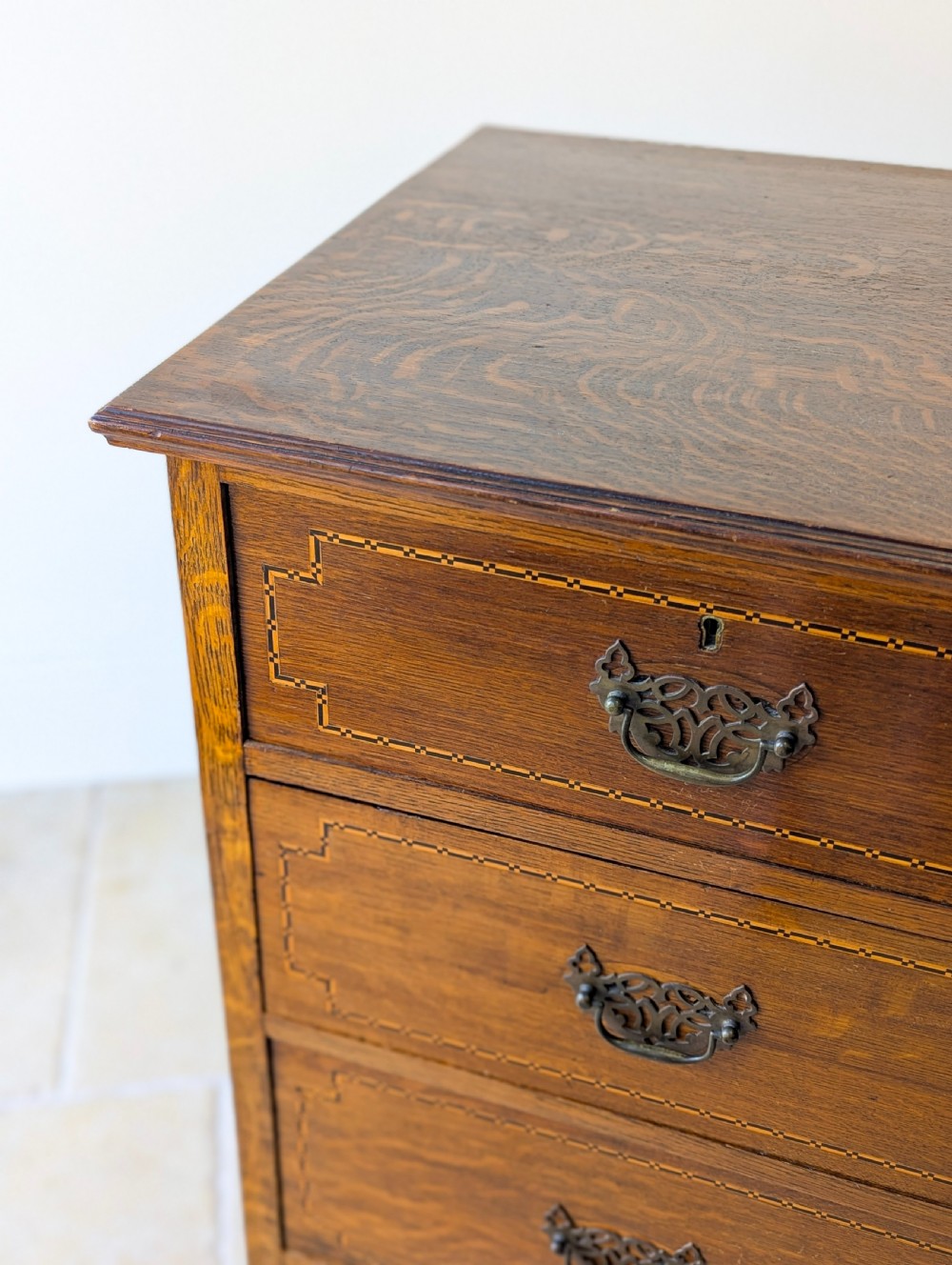 An Edwardian oak chest of drawers featuring a two over three layout with geometric boxwood inlay and dark metal drop handles on a plinth base. View II