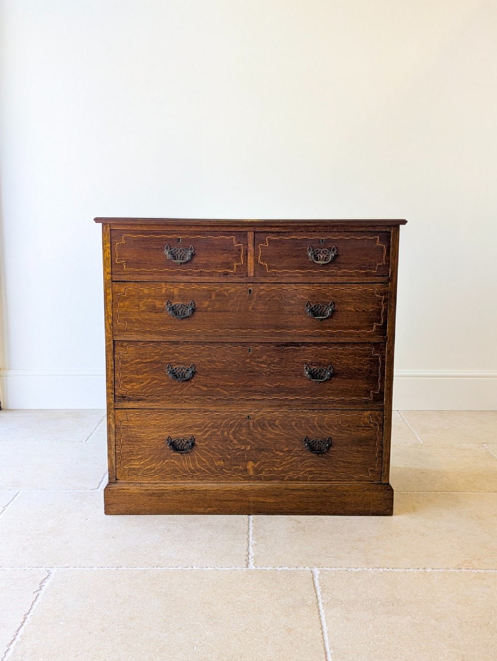 An Edwardian oak chest of drawers featuring a two over three layout with geometric boxwood inlay and dark metal drop handles on a plinth base. View III