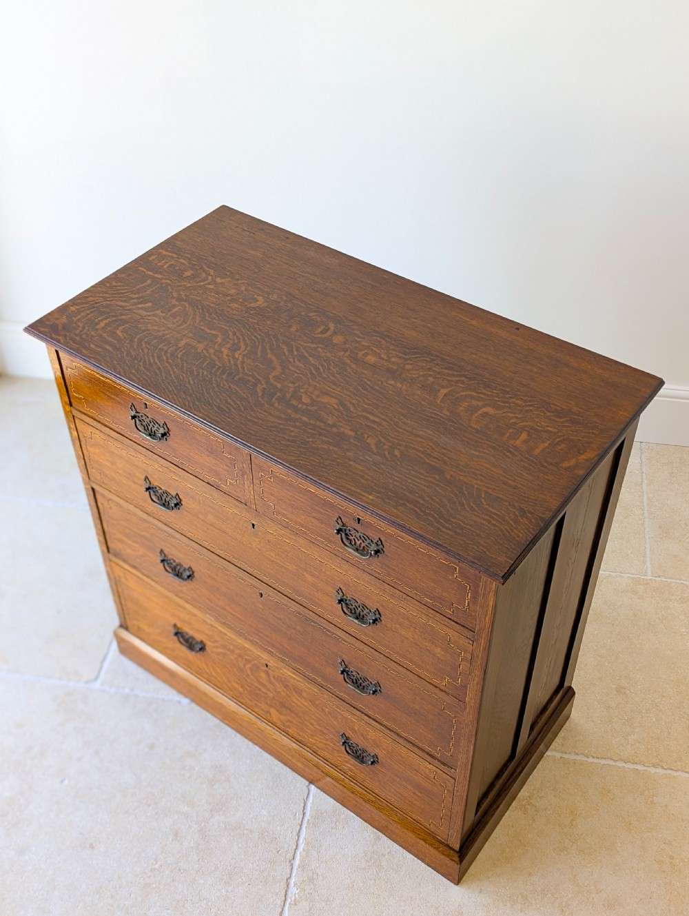 An Edwardian oak chest of drawers featuring a two over three layout with geometric boxwood inlay and dark metal drop handles on a plinth base. View IV