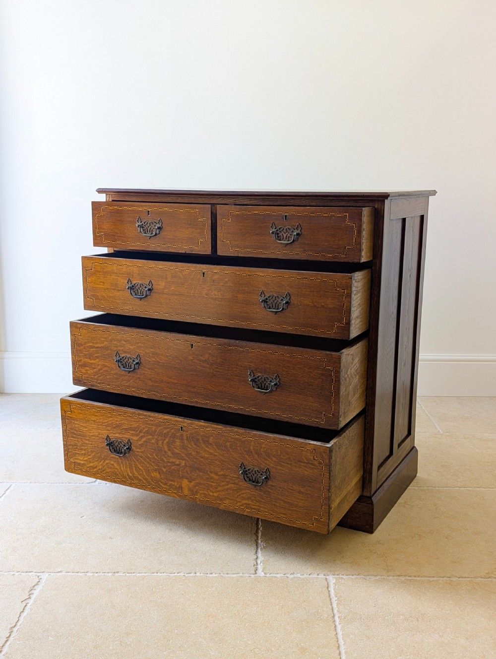 An Edwardian oak chest of drawers featuring a two over three layout with geometric boxwood inlay and dark metal drop handles on a plinth base. View V
