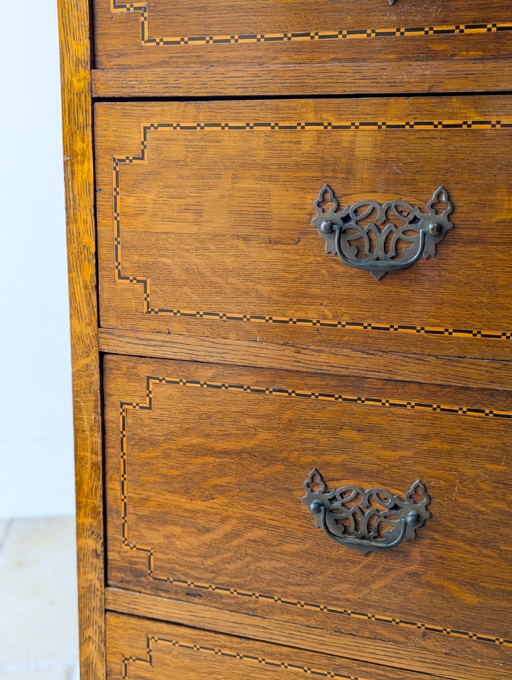 An Edwardian oak chest of drawers featuring a two over three layout with geometric boxwood inlay and dark metal drop handles on a plinth base. View VI