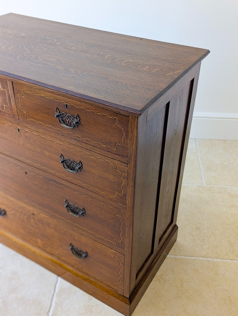 An Edwardian oak chest of drawers featuring a two over three layout with geometric boxwood inlay and dark metal drop handles on a plinth base. View VII