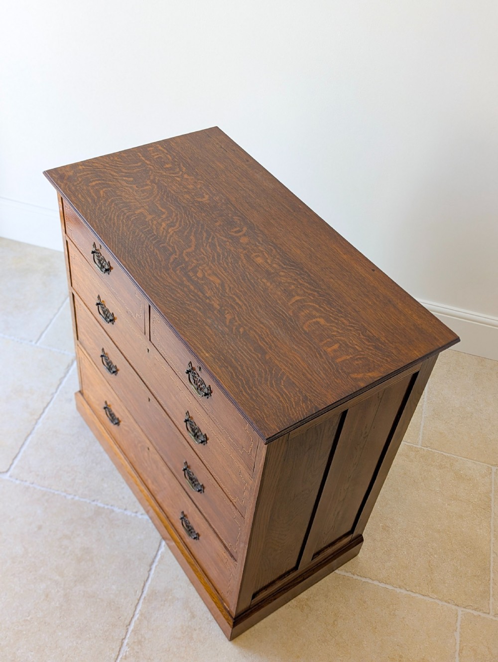 An Edwardian oak chest of drawers featuring a two over three layout with geometric boxwood inlay and dark metal drop handles on a plinth base. View VIII