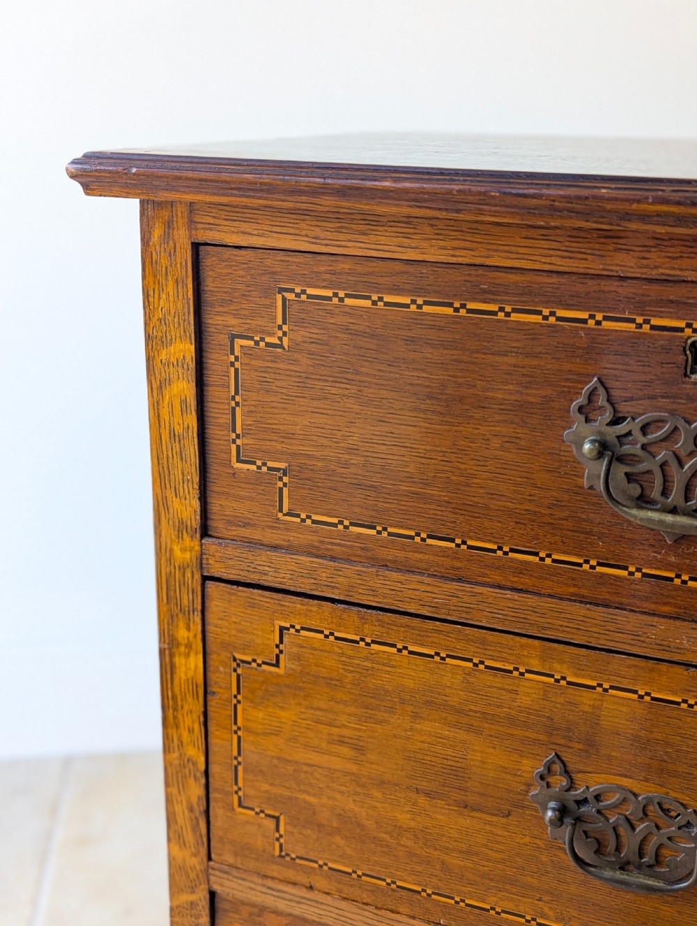 An Edwardian oak chest of drawers featuring a two over three layout with geometric boxwood inlay and dark metal drop handles on a plinth base. View IX