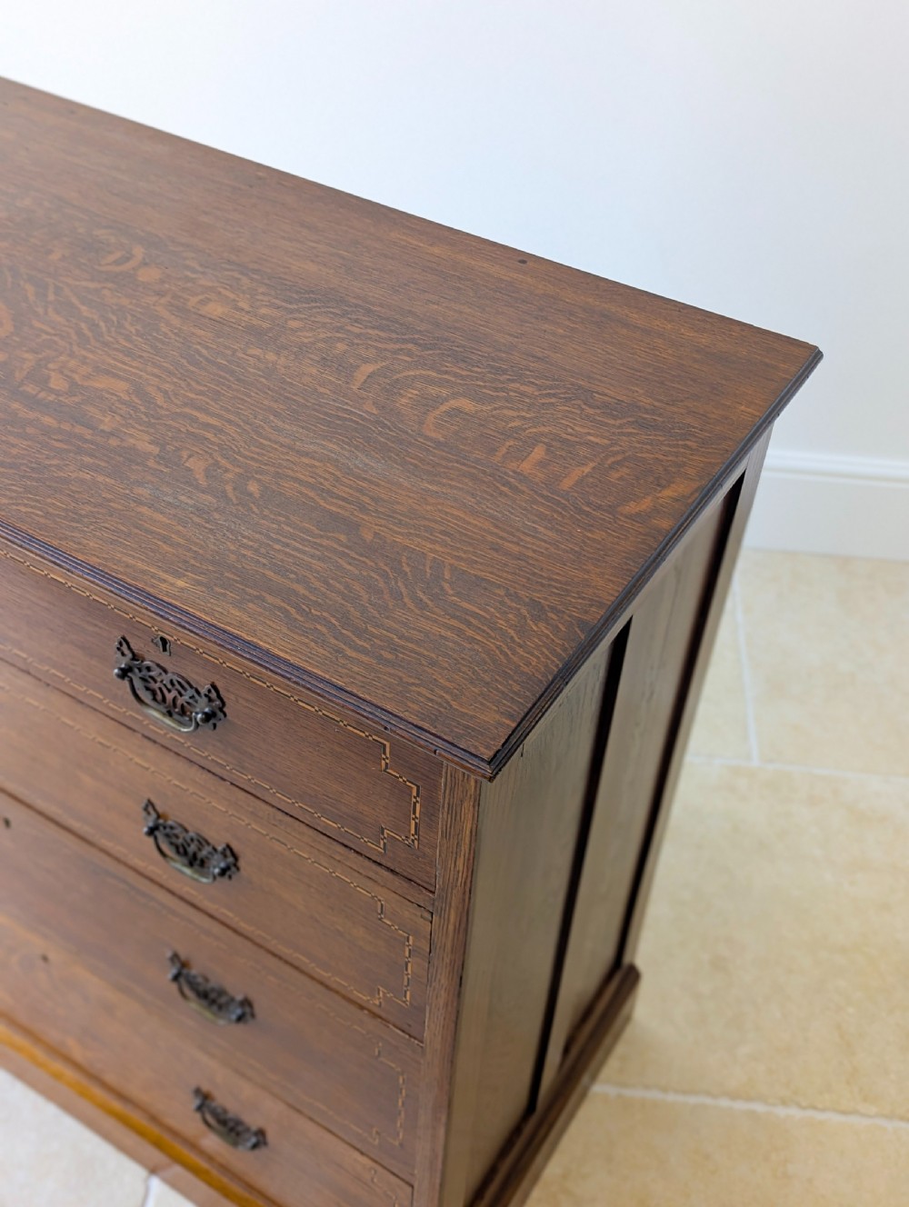 An Edwardian oak chest of drawers featuring a two over three layout with geometric boxwood inlay and dark metal drop handles on a plinth base. View X
