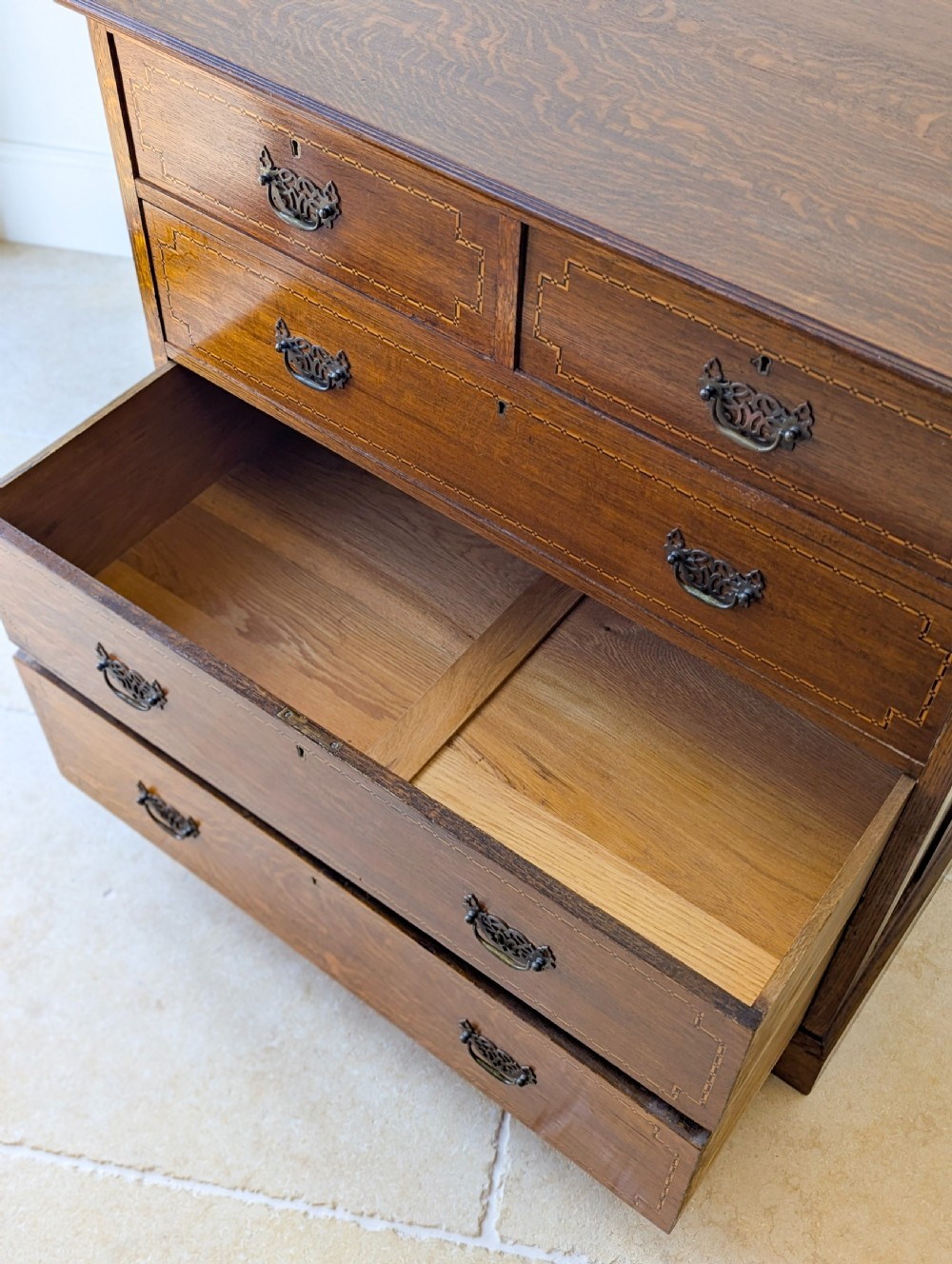 An Edwardian oak chest of drawers featuring a two over three layout with geometric boxwood inlay and dark metal drop handles on a plinth base. View XI