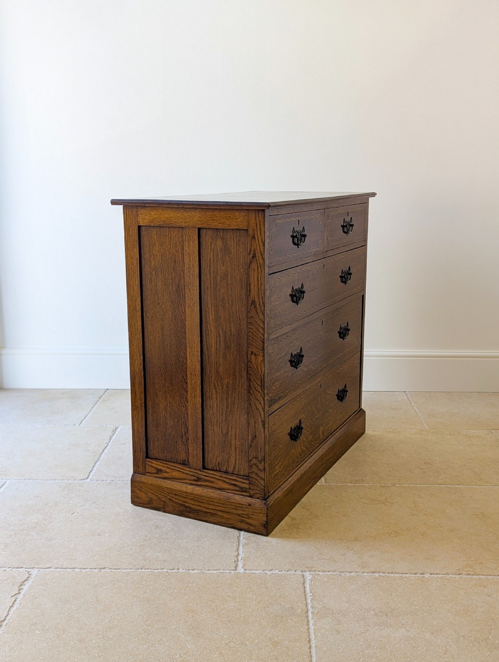 An Edwardian oak chest of drawers featuring a two over three layout with geometric boxwood inlay and dark metal drop handles on a plinth base. View XII