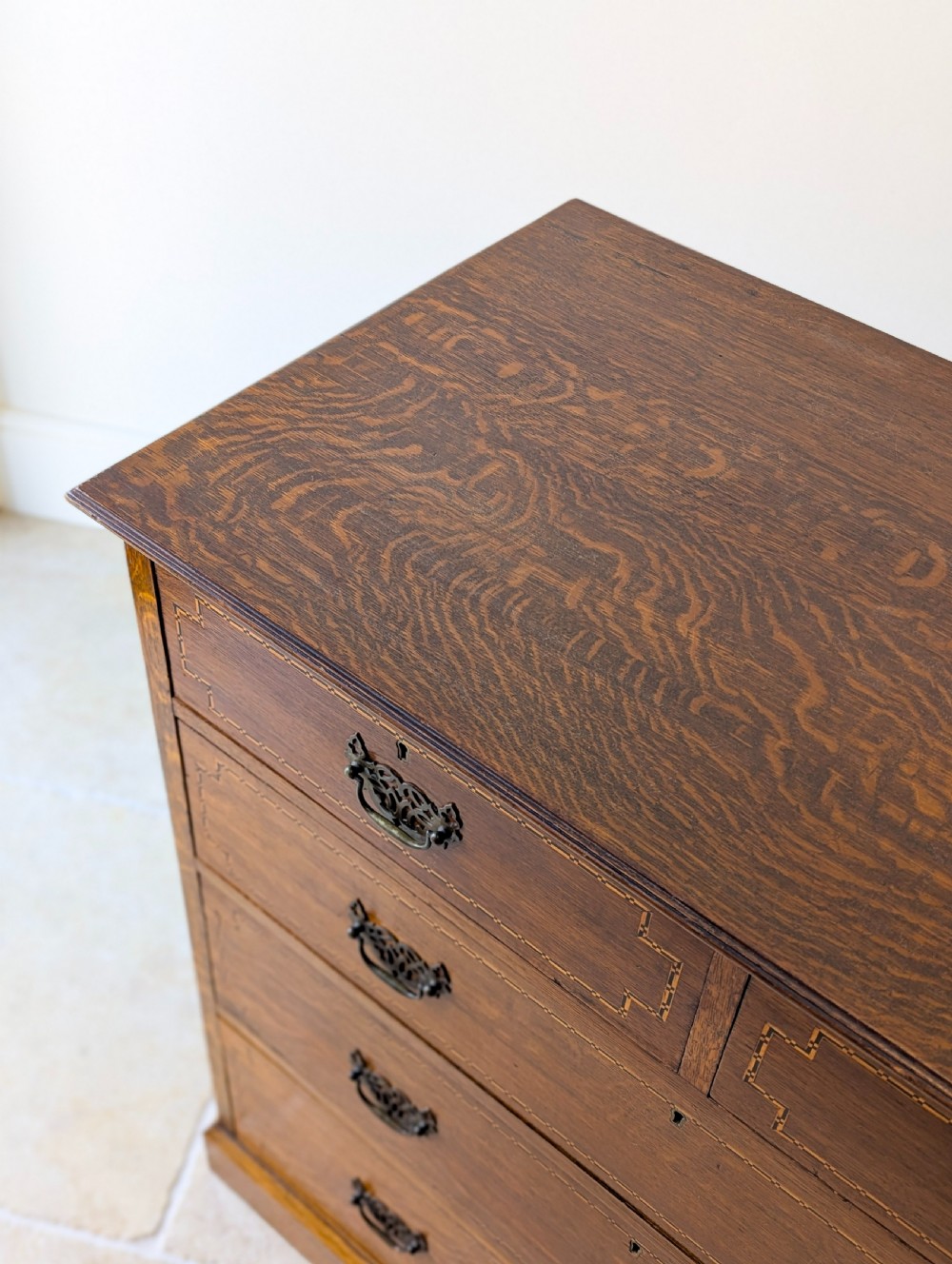 An Edwardian oak chest of drawers featuring a two over three layout with geometric boxwood inlay and dark metal drop handles on a plinth base. View XIII