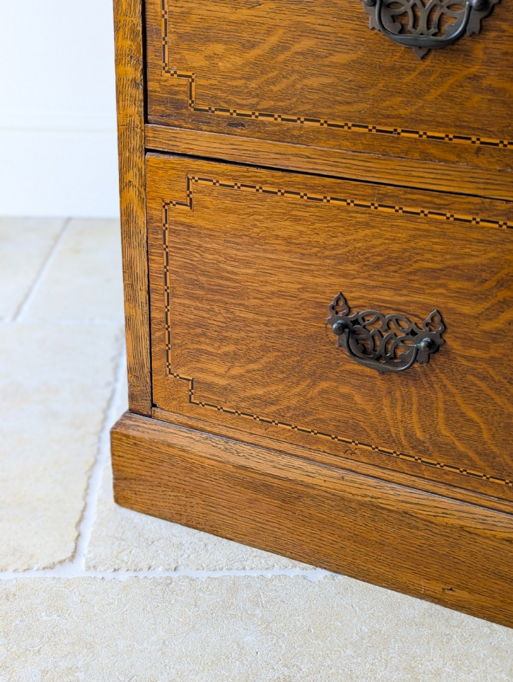 An Edwardian oak chest of drawers featuring a two over three layout with geometric boxwood inlay and dark metal drop handles on a plinth base. View XIV
