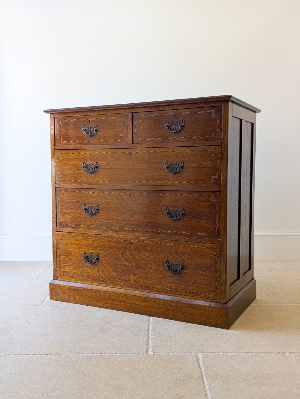 An Edwardian oak chest of drawers featuring a two over three layout with geometric boxwood inlay and dark metal drop handles on a plinth base. View XV