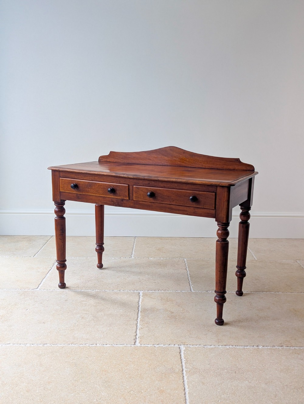 A Victorian mahogany writing table featuring two front drawers with dark turned knobs, a raised rear gallery upstand, and four turned legs.