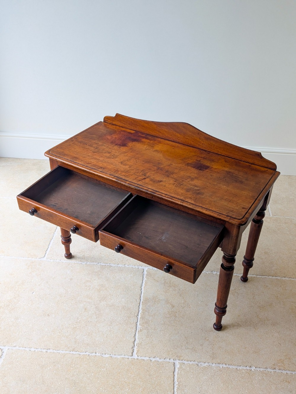 A Victorian mahogany writing table featuring two front drawers with dark turned knobs, a raised rear gallery upstand, and four turned legs. View IX