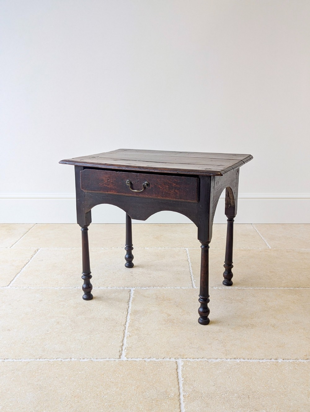 A dark oak Georgian lowboy featuring a single drawer with a brass drop handle, shaped frieze, and turned legs.