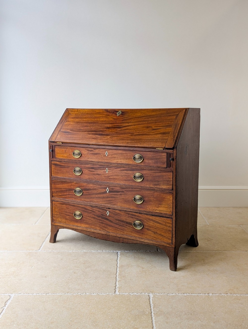 An antique Georgian mahogany slant-front bureau featuring four graduated long drawers with brass oval drop handles and shaped bracket feet.