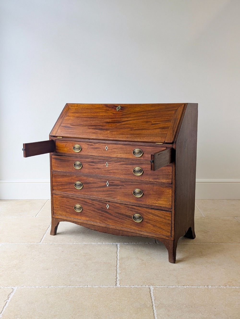 An antique Georgian mahogany slant-front bureau featuring four graduated long drawers with brass oval drop handles and shaped bracket feet. View V