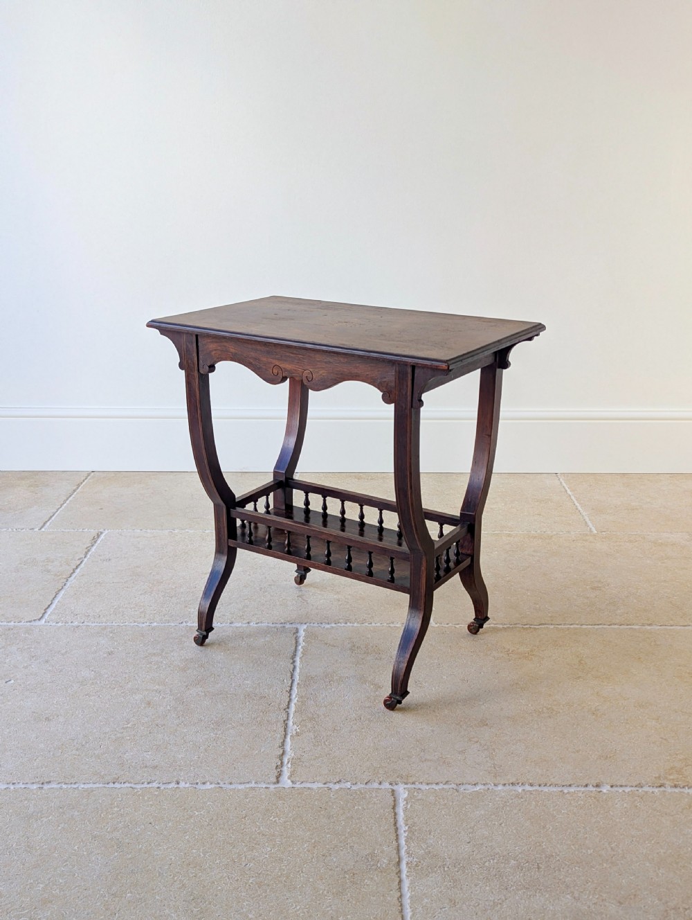 An antique Edwardian dark rosewood occasional table featuring out-swept legs, brass castors, a decorative shaped apron, and a lower shelf enclosed by turned vertical spindles.