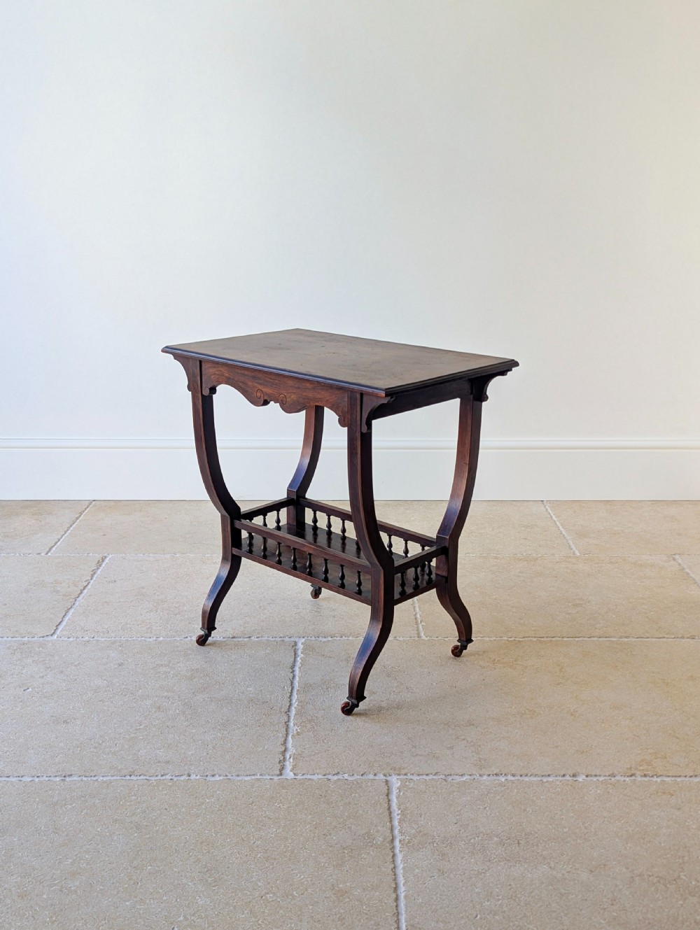 An antique Edwardian dark rosewood occasional table featuring out-swept legs, brass castors, a decorative shaped apron, and a lower shelf enclosed by turned vertical spindles. View V