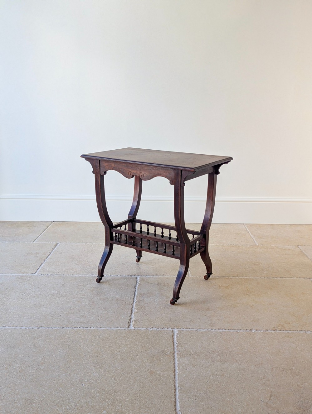 An antique Edwardian dark rosewood occasional table featuring out-swept legs, brass castors, a decorative shaped apron, and a lower shelf enclosed by turned vertical spindles. View VII