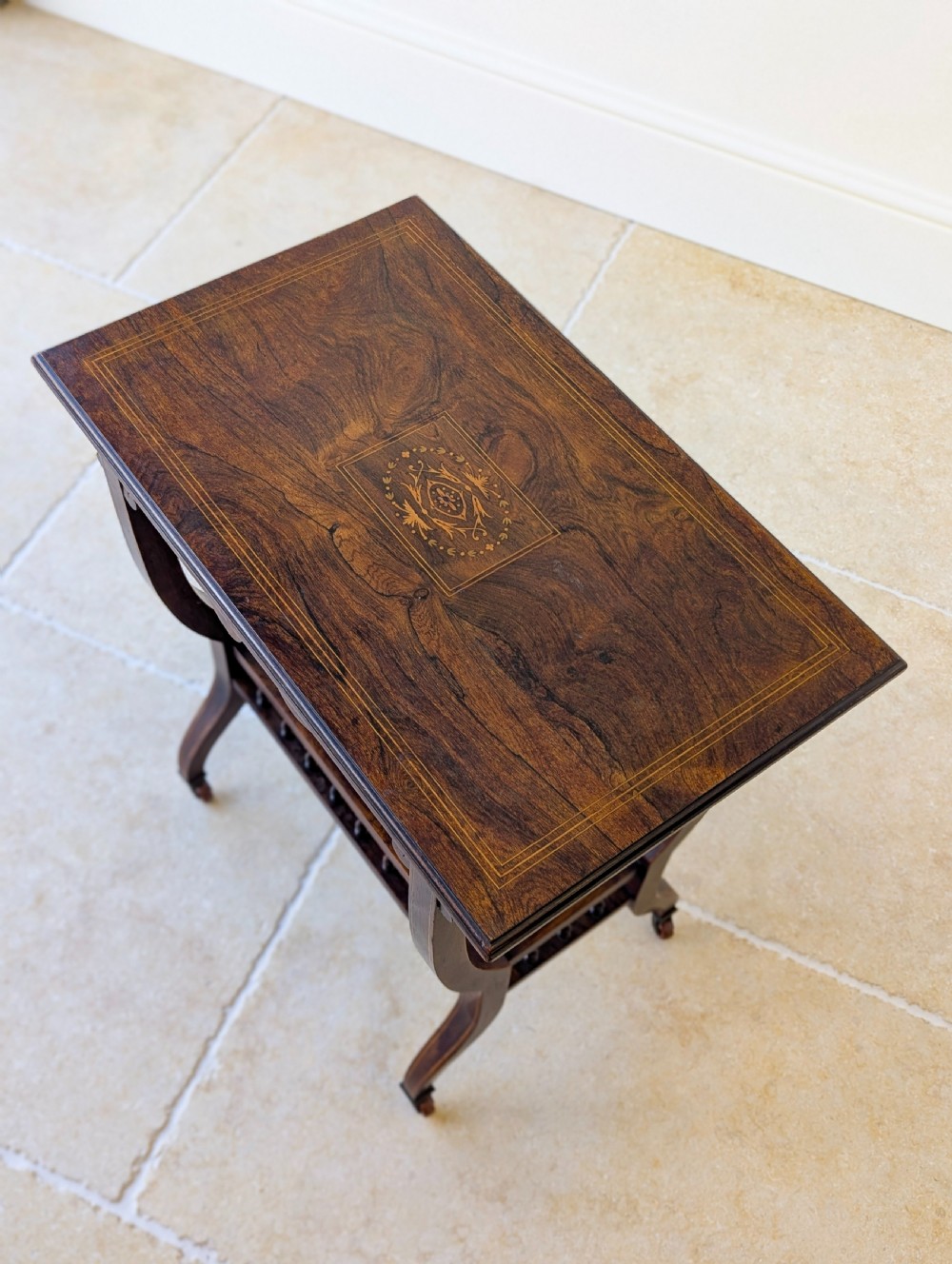 An antique Edwardian dark rosewood occasional table featuring out-swept legs, brass castors, a decorative shaped apron, and a lower shelf enclosed by turned vertical spindles. View X