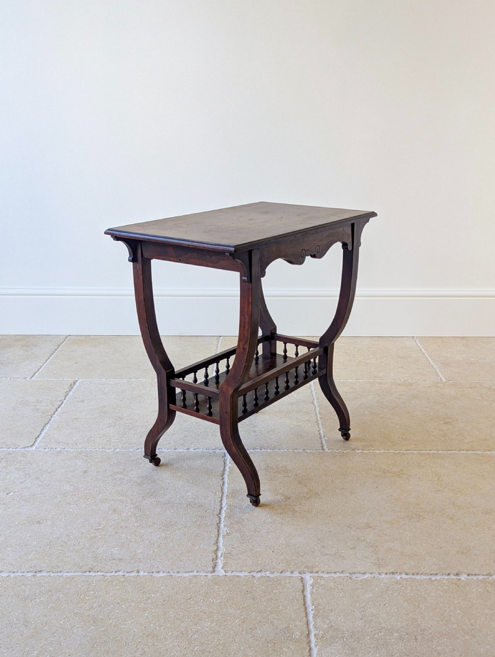 An antique Edwardian dark rosewood occasional table featuring out-swept legs, brass castors, a decorative shaped apron, and a lower shelf enclosed by turned vertical spindles. View XI