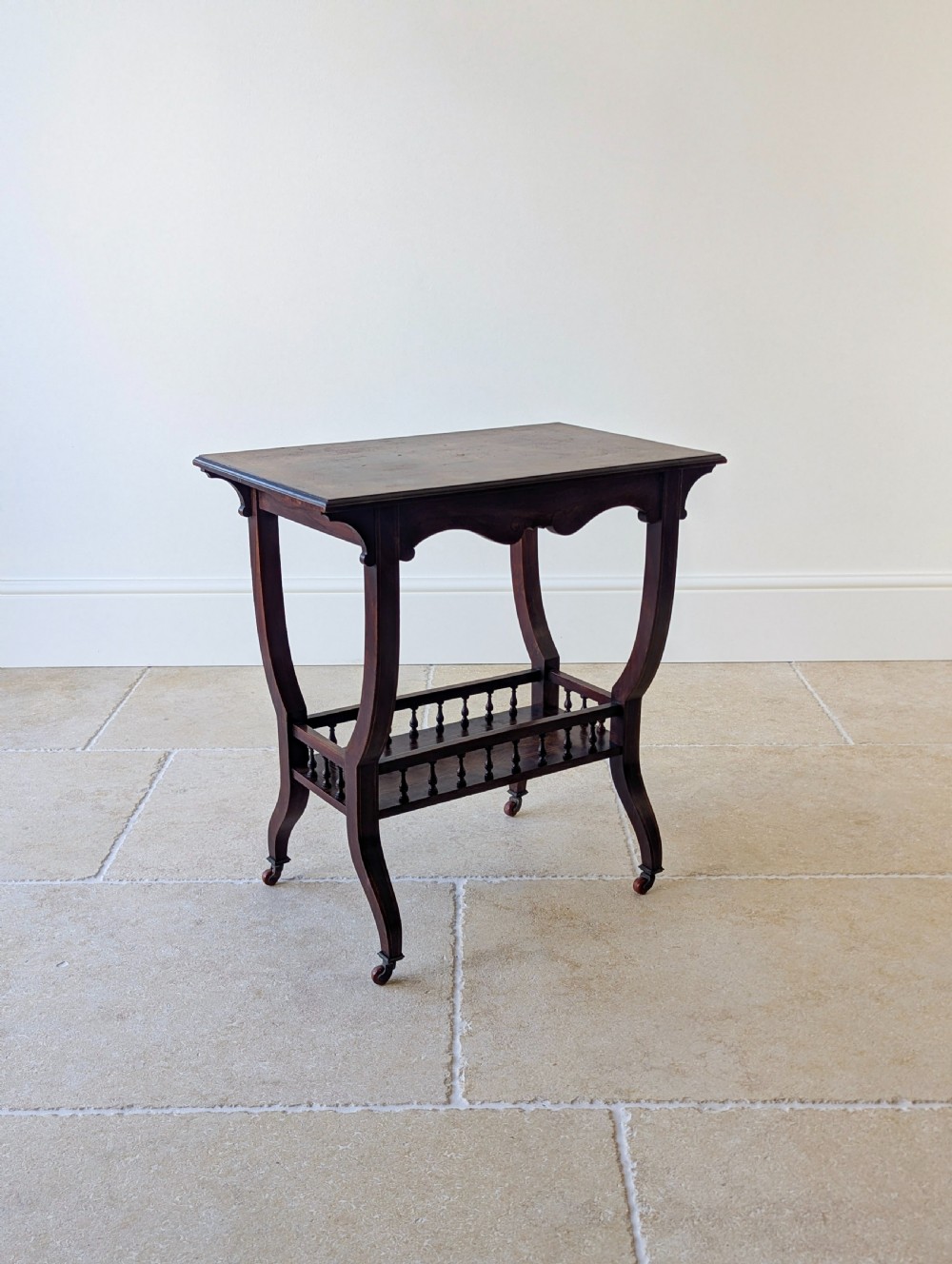 An antique Edwardian dark rosewood occasional table featuring out-swept legs, brass castors, a decorative shaped apron, and a lower shelf enclosed by turned vertical spindles. View XIV