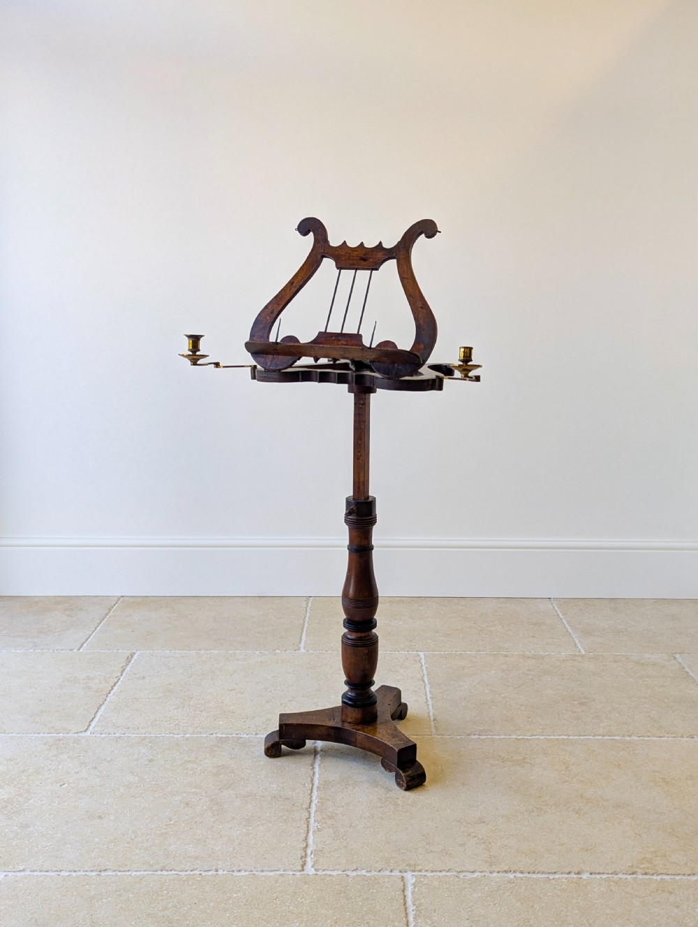An antique mahogany music stand featuring a lyre-shaped sheet rest, dual brass candle sconces, a turned stem, and a carved tripod base. View X