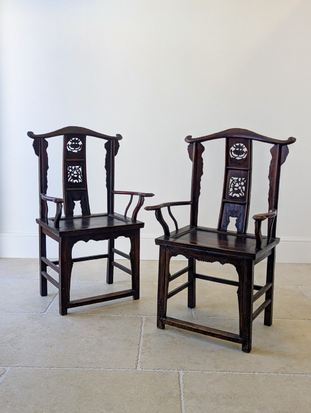 A pair of dark brown carved wooden Chinese yoke-back armchairs with curved top rails, open arms, and carved back splats.