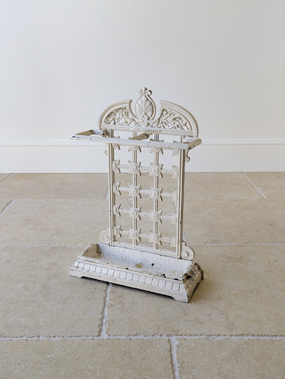 An antique off-white painted cast iron stick stand featuring an ornate backplate with shell motifs, a lattice panel, and a rectangular drip tray base.